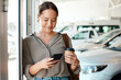 © J Bergen/peopleimages.com - Phone, coffee and woman at a car dealership typing a text message or scrolling on social media. Communication, technology and female person browsing on a mobile app with a cellphone in a showroom.