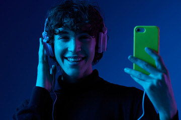 Wall Mural - Man smile with phone in hand looking at the smartphone screen in headphones listening to music, portrait dark blue background, neon light, style and trends, mixed light, men's fashion, copy space