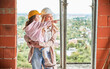 Woman in building helmet holding child while standing at construction site. Kid pointing away. Mother and daughter posing in apartment building under construction.