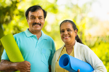 Wall Mural - portrait of Happy smiling indian senior couple with yoga mat looking at camera during morning workout at park - concept of fitness, active healthy lifestyle and wellness.