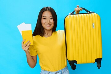 Wall Mural - Asian woman holding suitcase and passport with plane ticket smile with teeth in yellow t-shirt and blue jeans on blue background, copy space