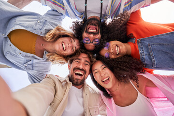 Selfie of multiethnic group of friends looking at camera cheerfully embracing each other. Happy group of people in circle looking down. Low angle view. Copy space.