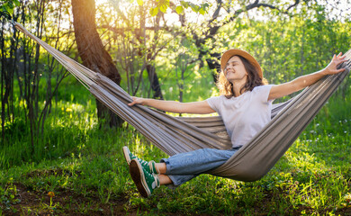 Wall Mural - Young happy caucasian woman in a hat lying in a hammock in a green garden enjoying a summer day