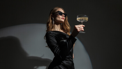 Beautiful elegant young fashion woman with sunglasses in a trendy black dress with a vintage glass of champagne on a dark background in the studio. Lady celebrating a birthday at a party