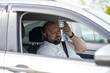 Weary overweight man drives car with broken air conditioner in hot summer weather. Male presses bottle of water to face to cool off suffering from heat, stuffiness. Exhausted tired overheated man.