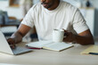 © Seventyfour - Close-up of African American man working online on laptop and drinking coffee while sitting at table in the kitchen