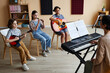© Seventyfour - Group of children sitting on chairs and playing musical instruments with teacher playing piano during lesson in music class