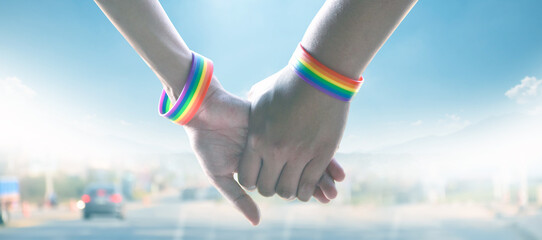 Closeup view of hands of gay couple wearing rainbow wristbands to present LGBT love, blurred rural road  and sunlight colors edited background, concept for LGBT people celebrations in pride month.