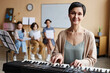 © Seventyfour - Portrait of mature music teacher smiling at camera while playing piano in music class with children in background