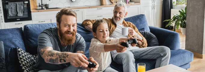 Wall Mural - KYIV, UKRAINE - APRIL 19, 2023: Positive gay man playing video game with daughter near partner and orange juice at home, banner