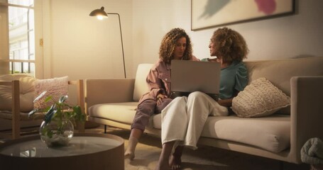 Wall Mural - Portrait of Two Brazilian Young Women Using Laptop Computer While Sitting on a Sofa at Home. Female Flatmates Doing Online Shopping Together, Checking Social Media, Enjoying The Evening in Living Room