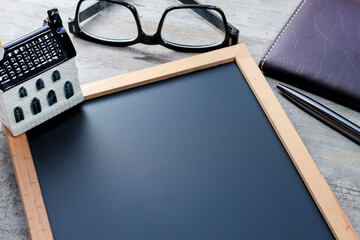 Black chalkboard on the wooden table with a house model, eyeglasses, pen, and notebook. Business and marketing concept