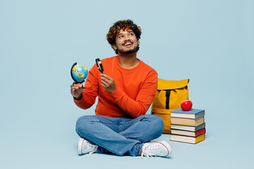 Wall Mural - Full body young teen Indian boy student wear casual clothes sit near books backpack bag hold Earth world globe use magnifier look aside isolated on plain blue background High school college concept