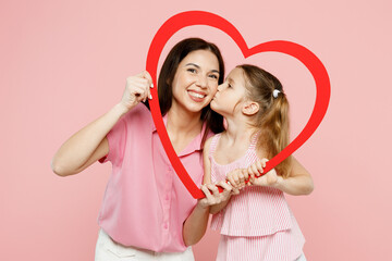 Wall Mural - Happy woman wear casual clothes with child kid girl 6-7 years old. Daughter kiss mother hold in hands look through big paper heart frame isolated on plain pink background. Family parent day concept.