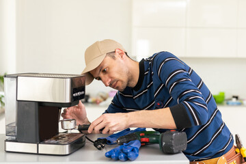 Wall Mural - Professional young repairman repairs a coffee maker. Handsome worker in uniform repairing coffee machine in a workshop.