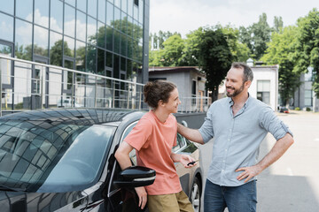 Wall Mural - happy bearded father looking at cheerful teenage son holding car key near automobile.