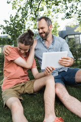 Sticker - joyful dad looking at teenager son and digital tablet while sitting in green park.