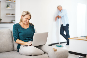 Poster - Its his day to do the housework. a mature woman using a laptop at home while her husband sweeps in the background.