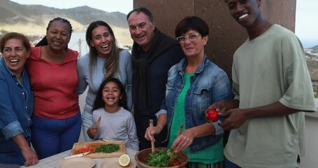 Wall Mural - Multiracial friends having fun smiling in front of camera while preparing food with child during weekend day - Multi generational people doing barbecue at home's rooftop while smiling on camera