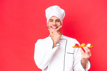 Wall Mural - Young caucasian chef man holding sashimi isolated on red background happy and smiling