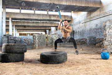 Shirtless muscular handsome man doing gym hammer tire smash explosively hitting his target hundreds and thousands of times in series, workout inside a industrial complex