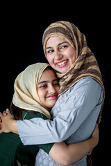 Wall Mural - Happy smiling Muslim girl with beautiful eyes wear hijab scarf and traditional costume hugging with her mother, warm love in family of mom and daughter, lovely Islamic family on dark black background.