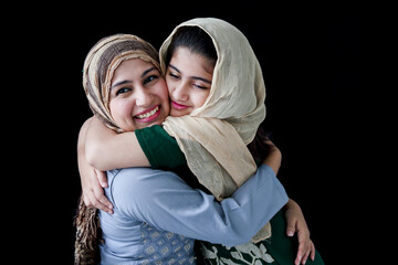 Wall Mural - Happy smiling Muslim girl with beautiful eyes wear hijab scarf and traditional costume hugging with her mother, warm love in family of mom and daughter, lovely Islamic family on dark black background.