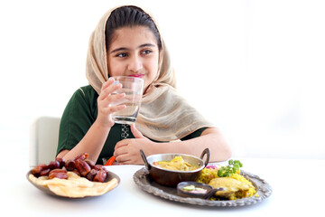 Wall Mural - Adorable Pakistani Muslim girl sitting at kitchen table with traditional Islamic halal food, kid with hijab drinks water, enjoy eating traditional Islamic halal food with hand on white background.