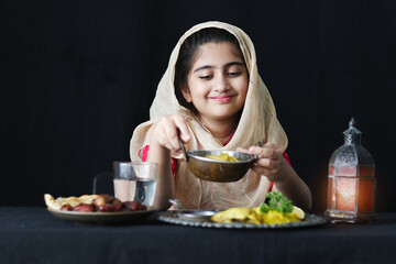 Wall Mural - Adorable happy smiling Muslim girl with beautiful eyes wearing traditional hijab, sitting at kitchen table, portrait of kid holding tray of Islamic halal food meal on dark black background.