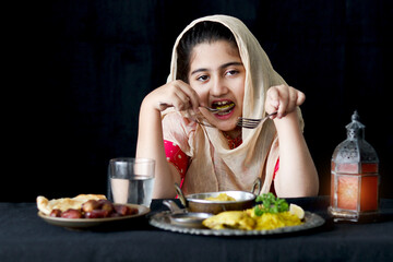 Wall Mural - Adorable happy Muslim girl with beautiful eyes wear hijab and traditional costume sit at kitchen table, portrait of kid eating Islamic halal food meal with spoon and fork on dark black background.