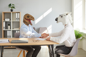 Happy funny colleagues business men shaking hands at meeting wearing animal masks sitting on their workplace at the office, celebrating success, making a deal or business achievement.