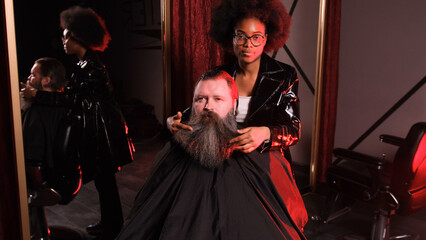 an African-American hairdresser girl straightens her beard with her hands to a male client sitting in an armchair.