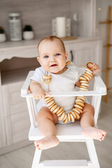 Wall Mural - a happy baby in a white cotton bodysuit with dryers or bagels around his neck on a high chair in a bright kitchen wants to eat, a small smiling baby boy or girl