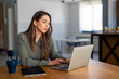 © Dorde - Serious millennial woman using laptop sitting at the table in a home office, typing, communicating online, writing emails, distantly working or studying on computer at home.