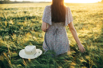 Wall Mural - Beautiful woman in floral dress walking in barley field in sunset light. Atmospheric tranquil moment, rustic slow life. Stylish female holding straw hat and enjoying evening summer countryside