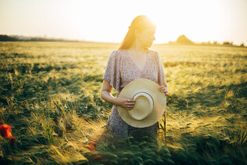 Wall Mural - Beautiful woman in floral dress standing in barley field in sunset light. Stylish female holding straw hat and relaxing in evening summer countryside. Atmospheric moment, rustic slow life