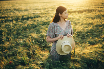 Wall Mural - Beautiful woman in floral dress standing in barley field in sunset light. Atmospheric tranquil moment, rustic slow life. Stylish female holding straw hat and enjoying evening summer countryside