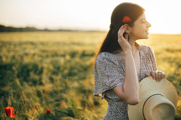 Wall Mural - Beautiful woman with poppy flower behind ear standing in sunset light in barley field. Atmospheric tranquil moment, rustic slow life. Stylish female enjoying evening summer countryside