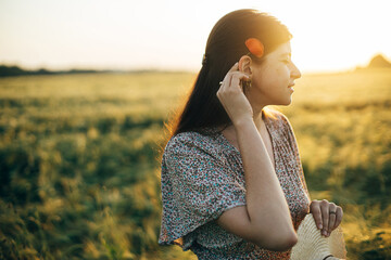 Wall Mural - Beautiful woman with poppy flower behind ear standing in sunset light in barley field. Atmospheric tranquil moment, rustic slow life. Stylish female enjoying evening summer countryside