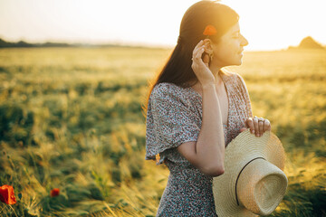 Wall Mural - Beautiful woman with poppy flower behind ear relaxing in barley field in sunset light. Stylish female standing in evening summer countryside. Atmospheric moment, rustic slow life