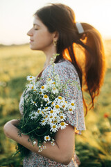 Wall Mural - Beautiful woman with wildflowers enjoying sunset in barley field. Atmospheric tranquil moment, rustic slow life. Stylish female gathering wildflowers and relaxing in evening summer countryside