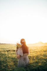 Wall Mural - Beautiful woman with wildflowers enjoying sunset in barley field. Atmospheric tranquil moment, rustic slow life. Stylish female gathering flowers and relaxing in evening summer countryside