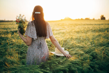 Wall Mural - Beautiful woman with wildflowers enjoying sunset in barley field. Atmospheric tranquil moment, rustic slow life. Stylish female gathering flowers and relaxing in evening summer countryside