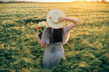 Wall Mural - Beautiful barley field in sunset light and blurred image of woman in straw hat holding wildflowers. Evening summer countryside and gathering flowers. Atmospheric tranquil moment