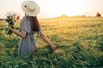 Wall Mural - Beautiful woman in hat with wildflowers enjoying sunset in barley field. Atmospheric tranquil moment, rustic slow life. Stylish female gathering flowers and relaxing in evening summer countryside