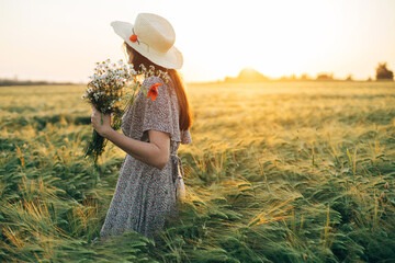 Wall Mural - Beautiful woman in hat with wildflowers enjoying sunset in barley field. Atmospheric tranquil moment, rustic slow life. Stylish female gathering flowers and relaxing in evening summer countryside