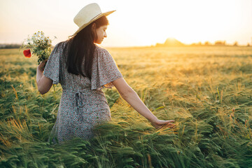 Wall Mural - Beautiful woman in hat with wildflowers bouquet standing in barley field in sunset light. Stylish female relaxing in evening summer countryside and gathering flowers. Atmospheric tranquil moment
