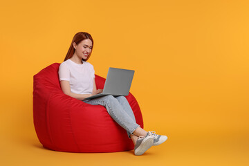 Smiling young woman working with laptop on beanbag chair against yellow background, space for text