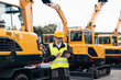 © scharfsinn86 - Engineer in a helmet with a digital tablet stands next to construction excavators