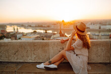 Beautiful Woman In Hat Enjoys Stunning View Of The City At Dawn. Back View. Tourist Is Looking At Landscape Of City From Height. Travel In Europe.
